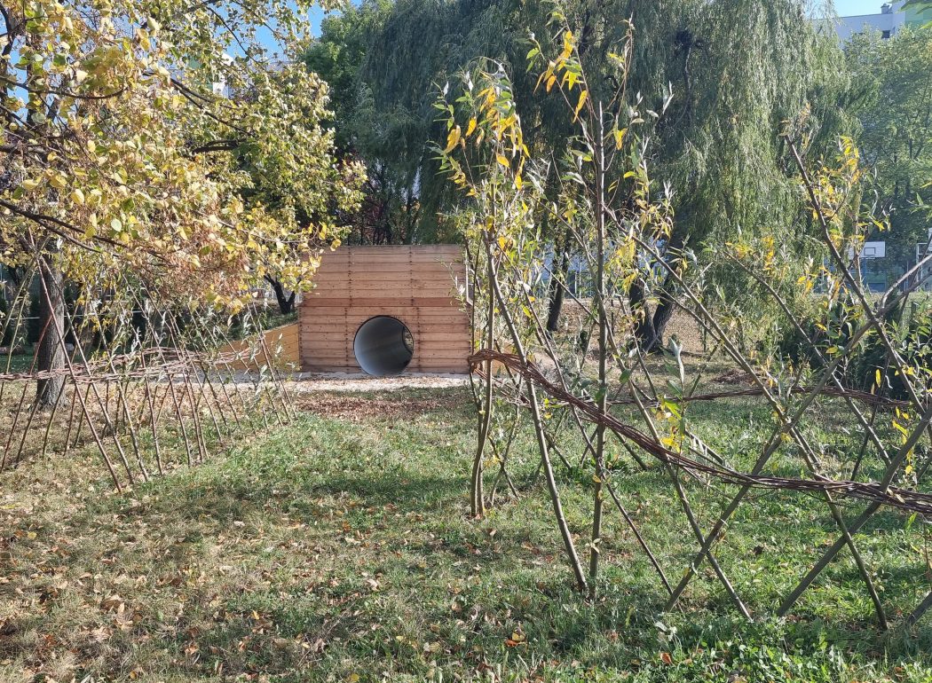 zielona trawa, wigwam z rosnących gałęzi, w oddali tunek z górką