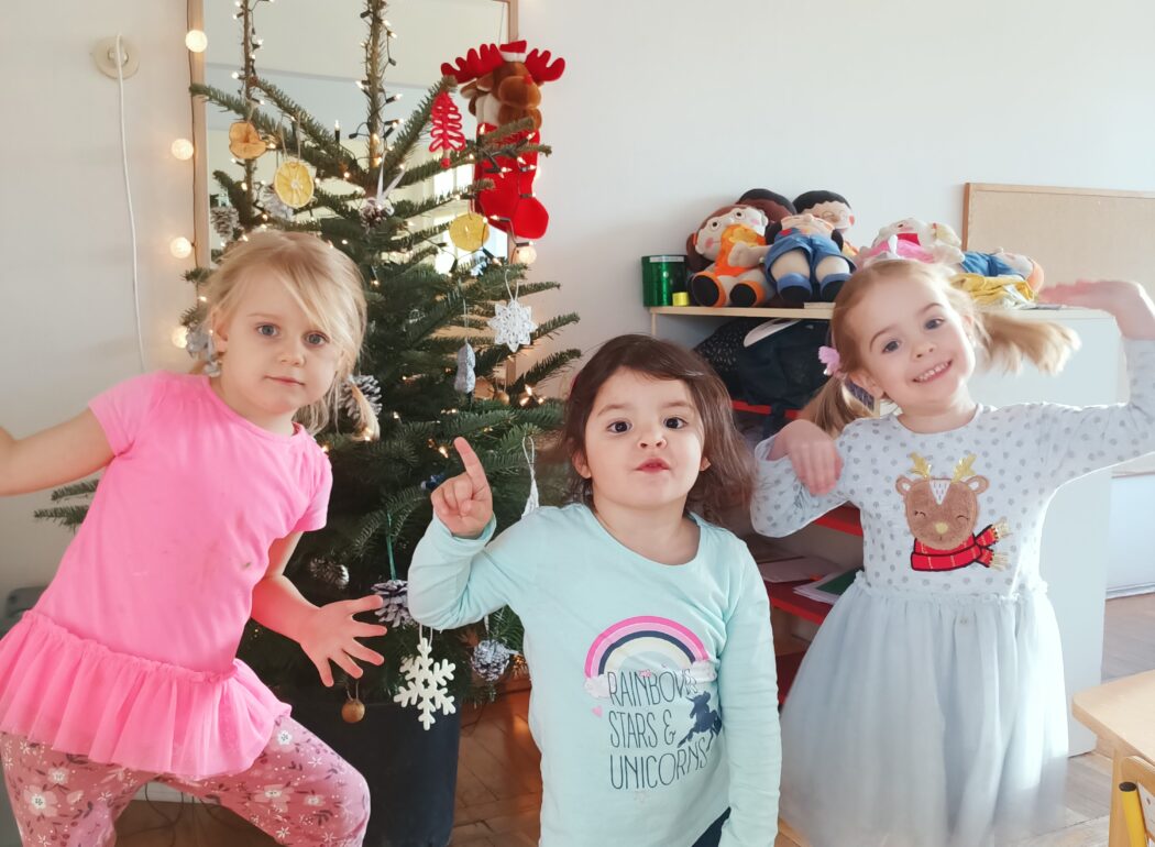 Trzy uśmiechniete dziewczynki stoją, za nimi udekorowana choinka
