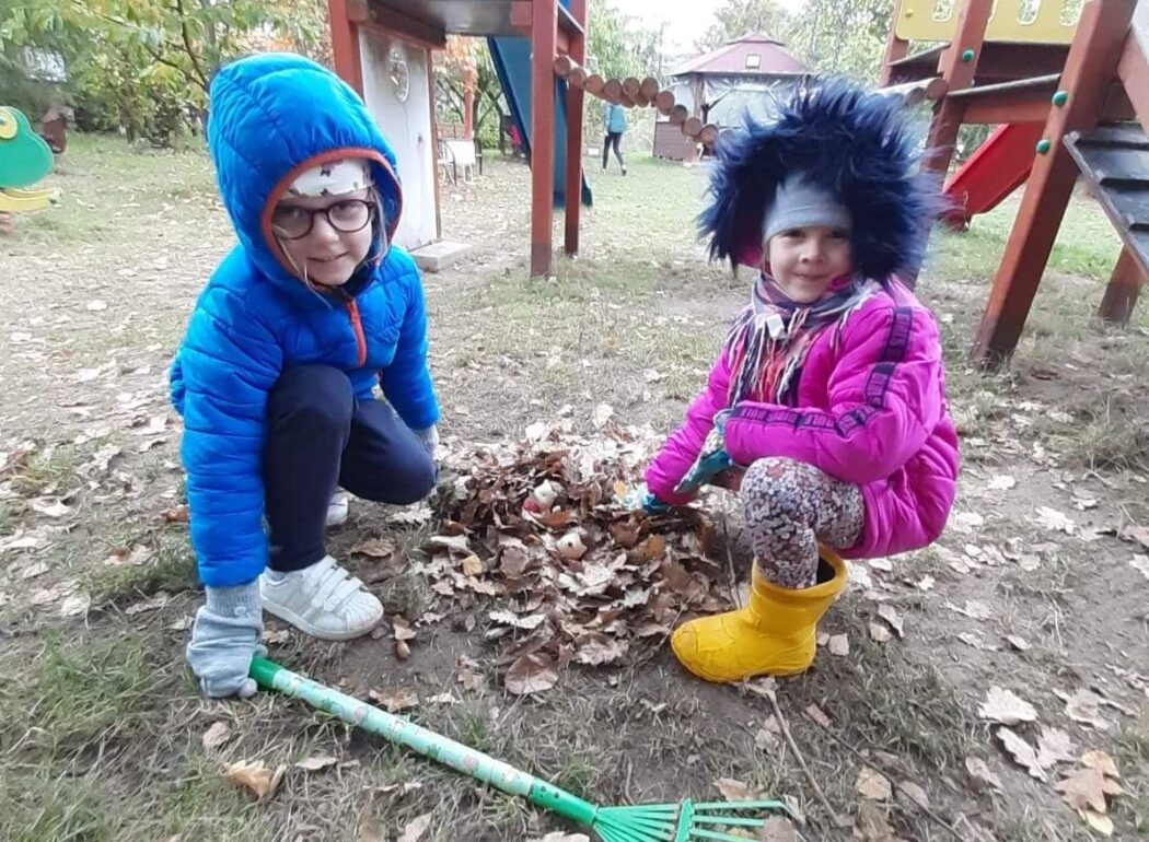 dwie dziewczynku kucają na ziemi koło sterty liści i grabi