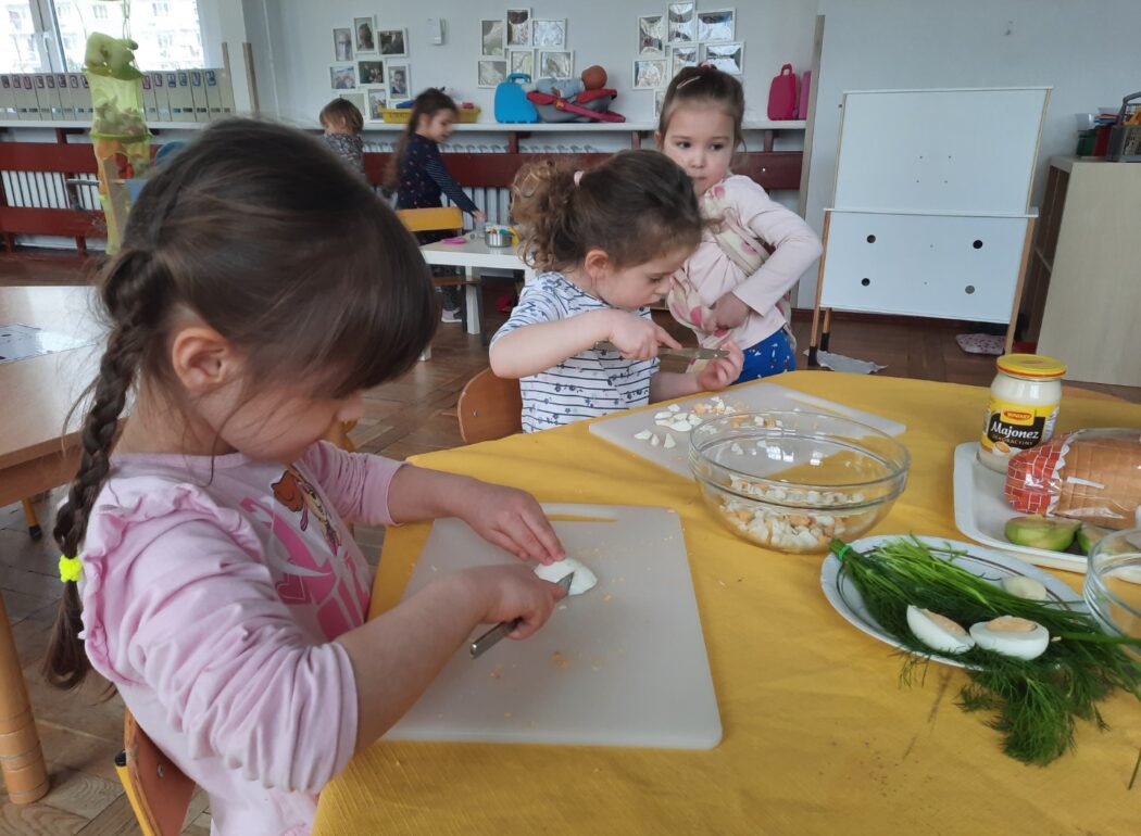 dzieci kroją jajka, na stole są miseczki z jajkiem pokrojonym, talerzyk z pietruszką zieloną i koperkiem