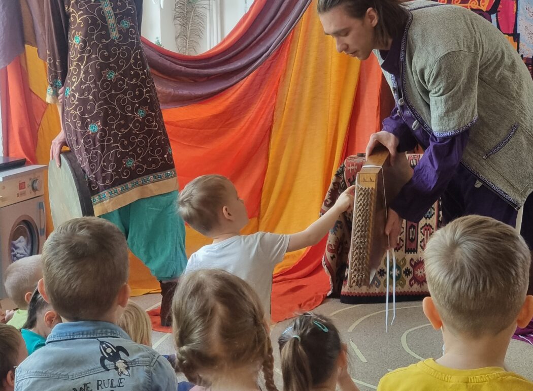 mężczyzna pochylającyc się nad dzieckiem z instrumentem ludowym w tle uśmiechająca się kobieta