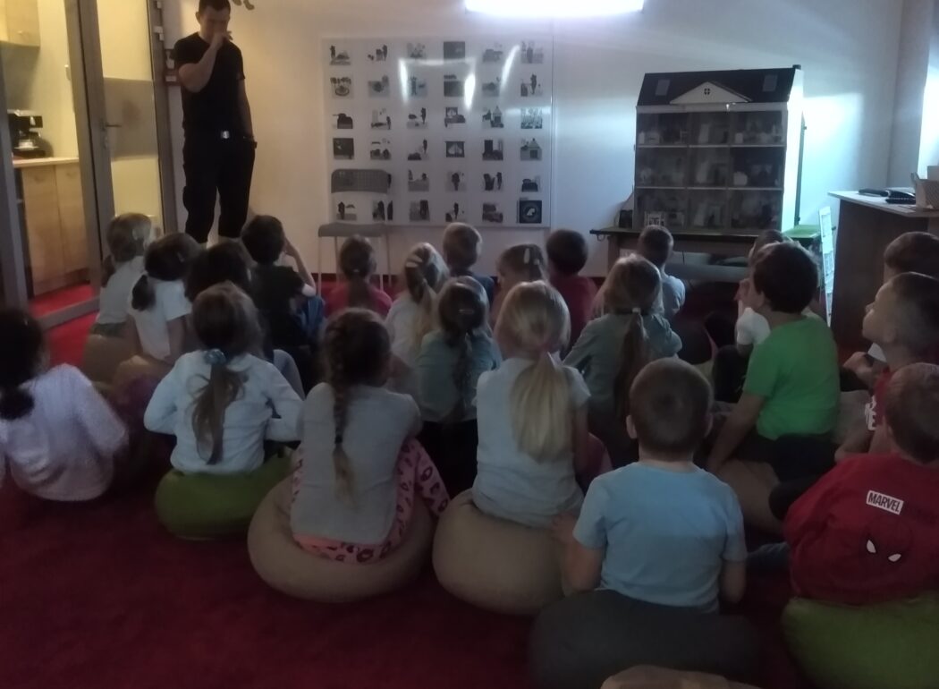 w ciemnej sali dzieci siedzą na dywanie a na końcu sali stoi strażak