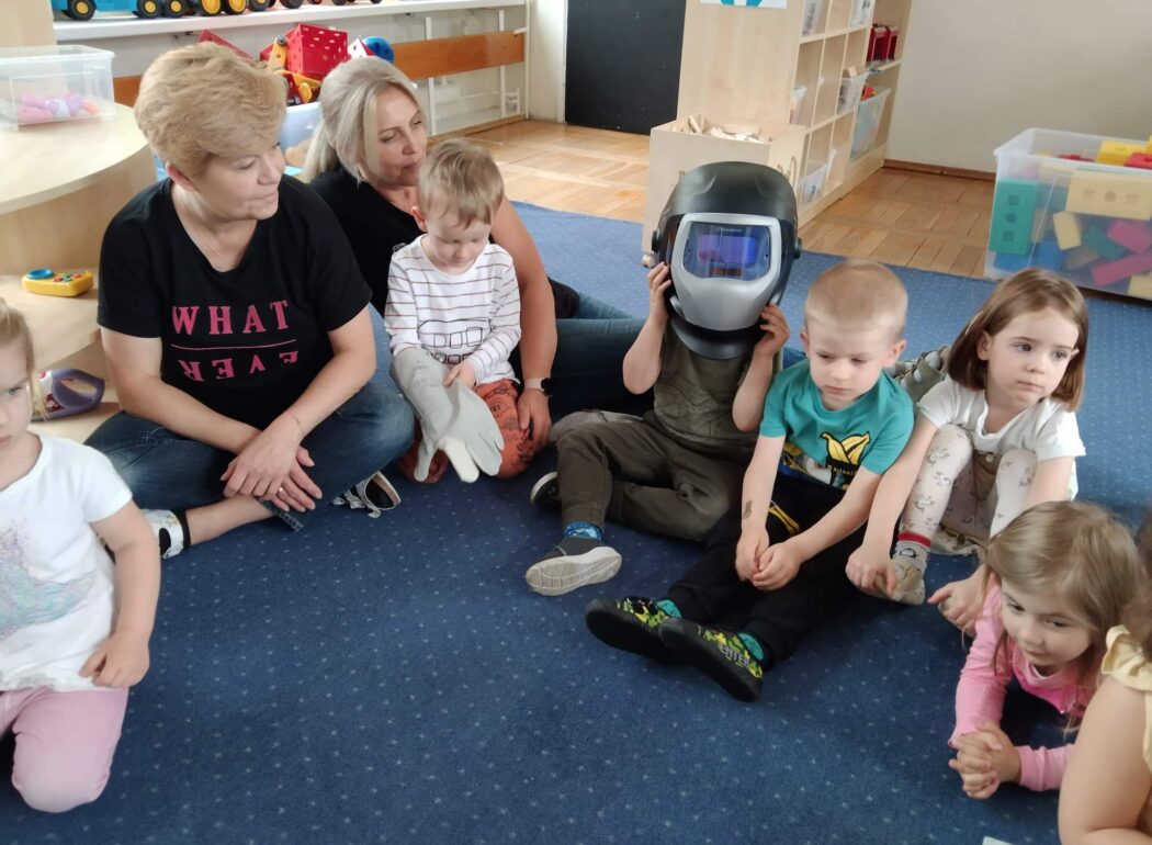 dzieci siedzą na niebieskim dywanie, jedno ma kask na głowie
