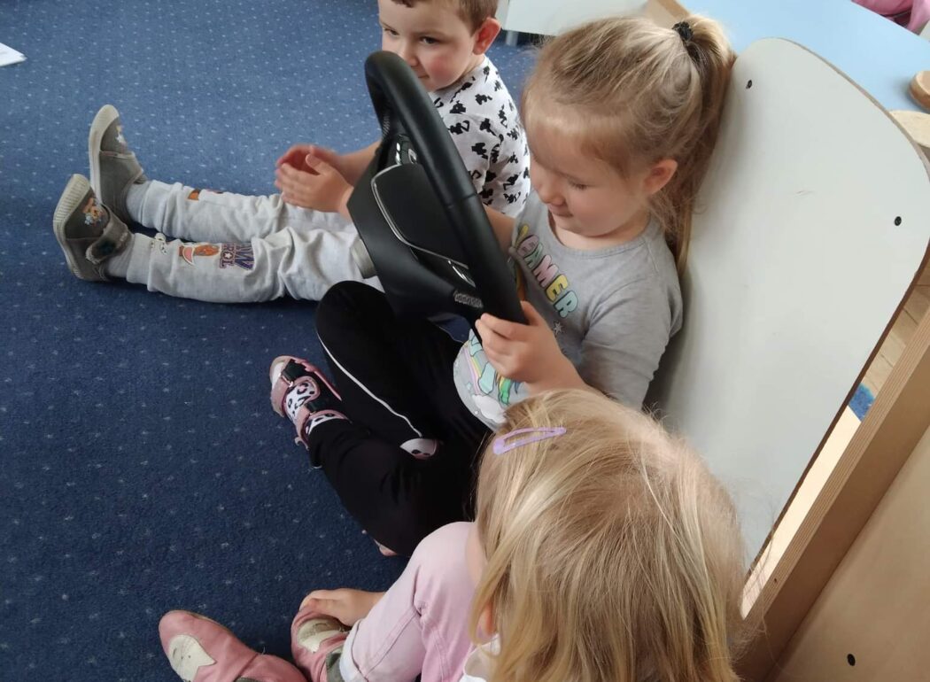 dzieci siedzą na dywanie, dziewczyna trzyma w ręku samochodową kierownicę