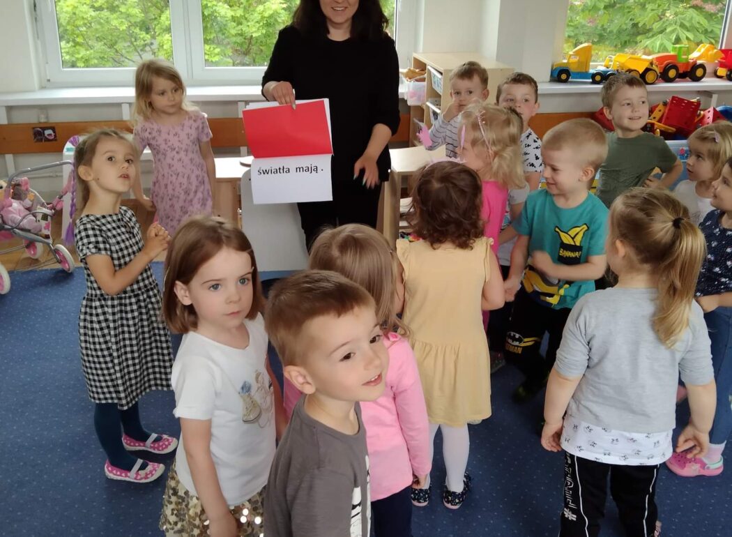 grupka dzieci stoi na dywanie, na srodku stoi kobieta trzymająca w ręku czerwoną kartkę z napisem u dołu światła mają