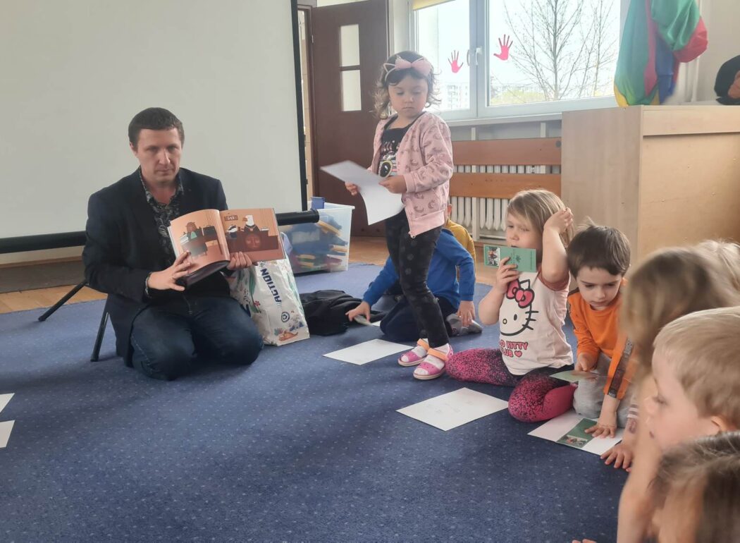 mężczyzna trzymający w rękach książeczkę po jego prawej stronie dzieci siedzące na niebieskim dywanie