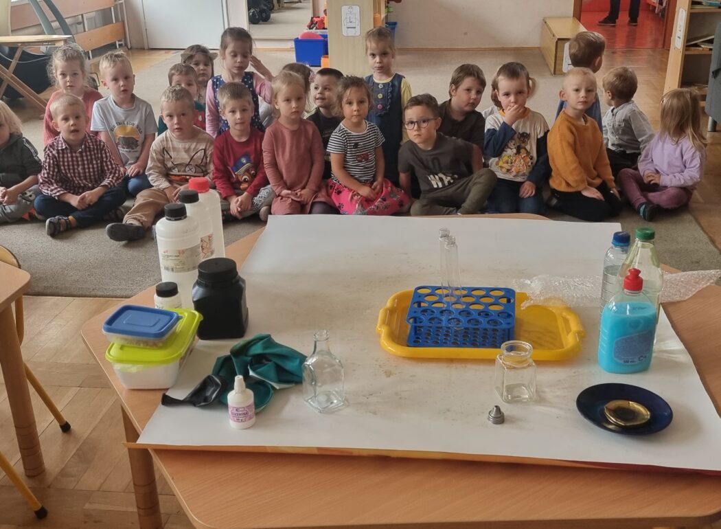 na stoliku stoją buteleczki, menzurki, środki chemiczne w buteleczkach i pudełkach, za stolikiem na dywanie siedzą dzieci