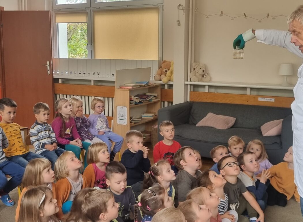 mężczyzna w białym kitlu i rękawiczkach trzyma w ręku pojemnik z płynem, przed nim na dywanie siedzą dzieci