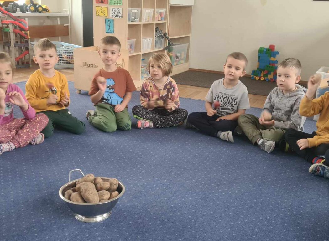 dzieci siedzą ze skrzyżowanymi nogami na niebieskim dywanie, przed nimi stoi miska ziemniakami