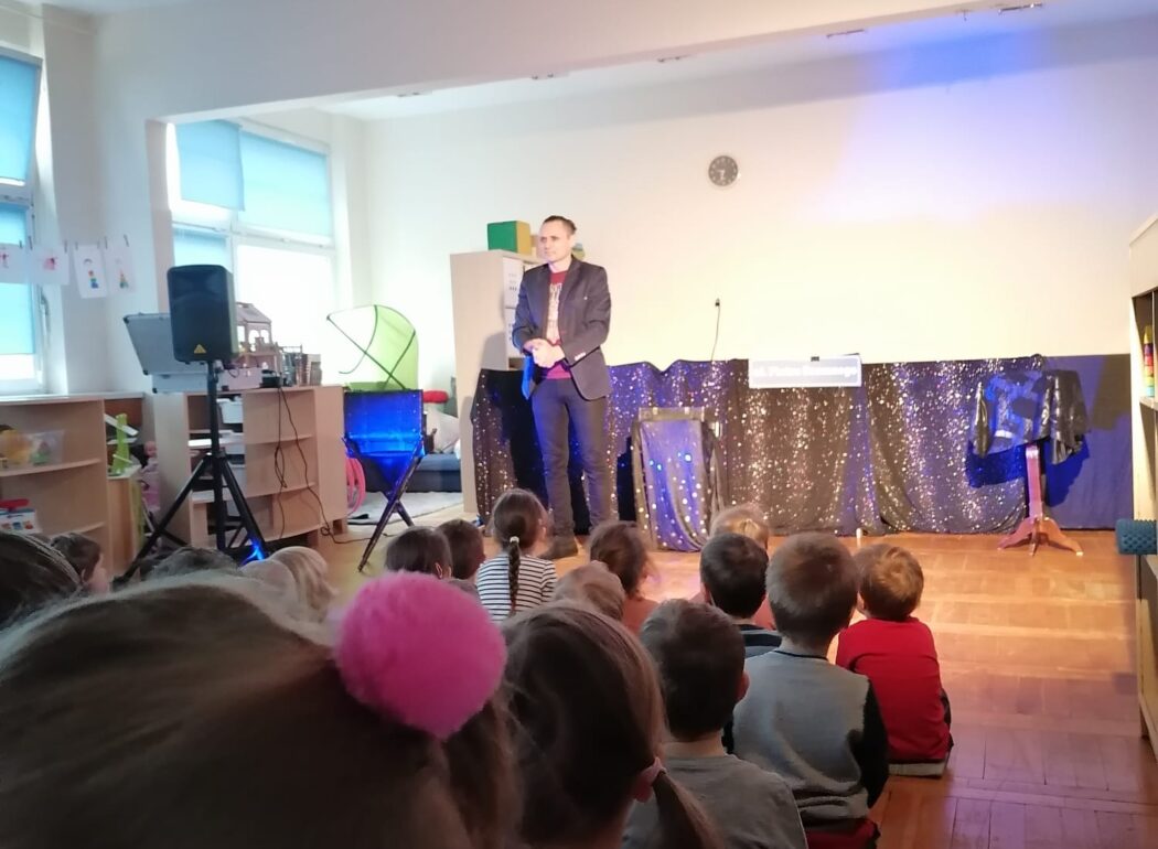 dzieci siedzą na podłodze zwrócone strone podświetlonej sceny na której stoi magik