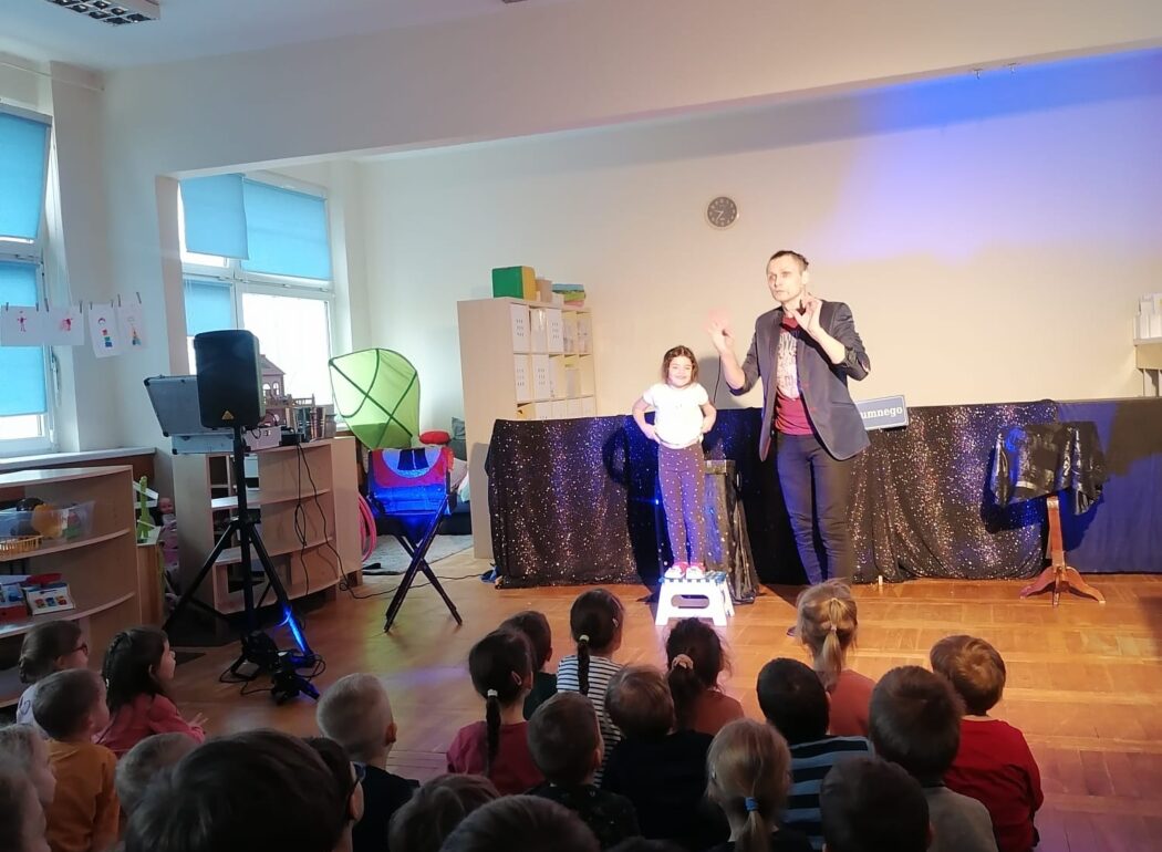 dzieci siedzą na podłodze zwrócone w stronę podświetlonej sceny na której stoi magik i dziewczynka