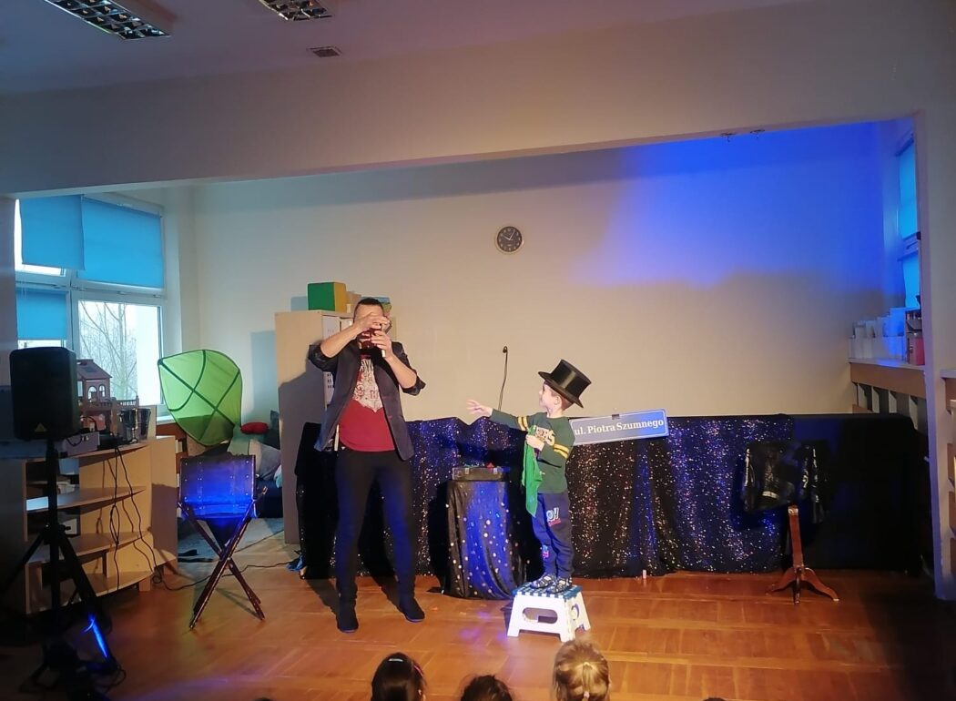 dzieci siedzą na podłodze zwrócone w stronę podświetlonej sceny na której stoi magik i chłopiec w czarnym cylindrze