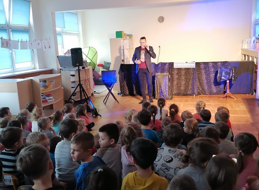 dzieci siedzą na dywanie zwrócone w stronę podświetlonej sceny, na której stoi magik