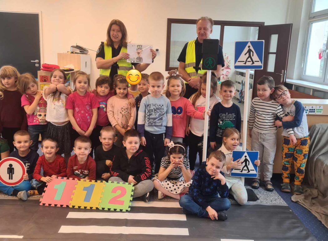 dzieci ustawione do zdjęcia trzymające znaki drogowe i puzzle z cyframi jeden jeden dwa, za nimi pan i pani ze straży miejskiej w odblaskowych kamizelkach