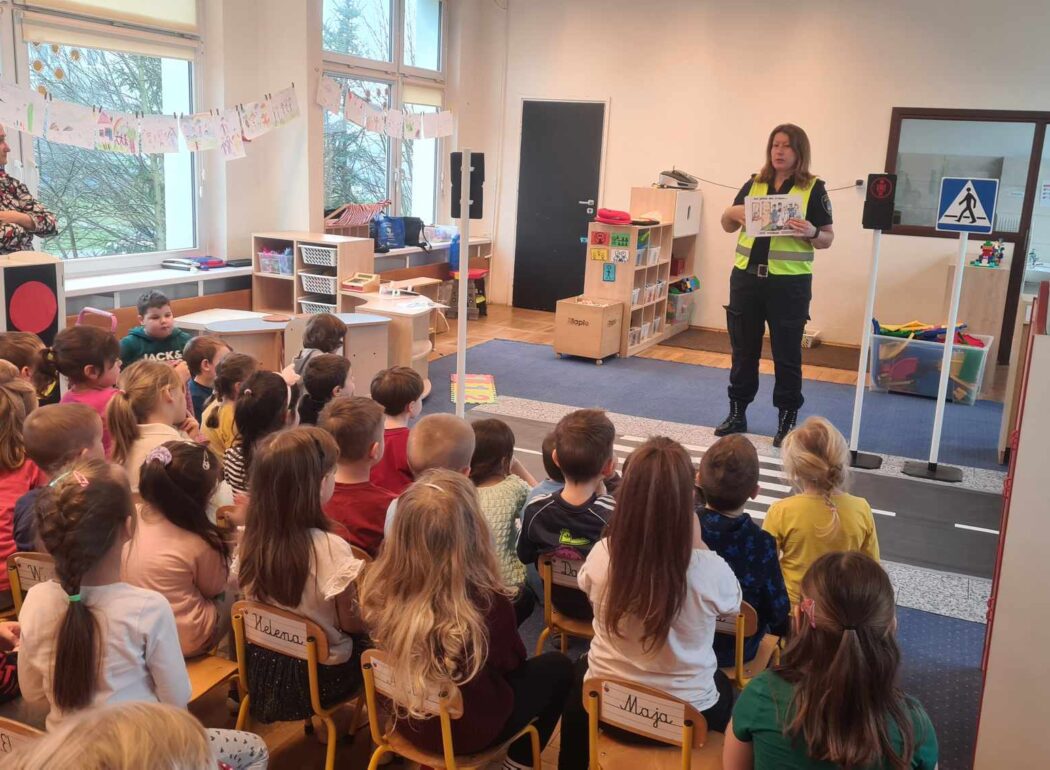 dzieci siedzą na krzesełkach i patrzą na panią ze straży miejskiej trzymającą kartkę z obrazkiem