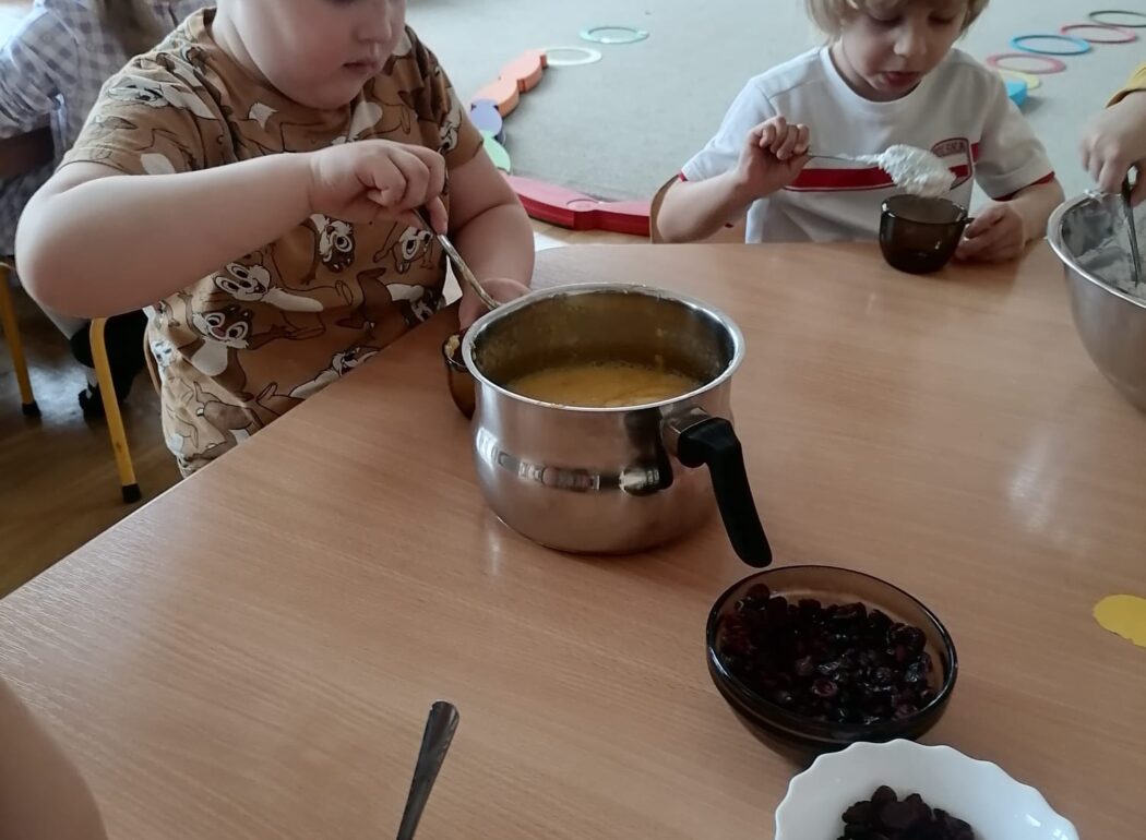 chłopcy siedząc przy stole nakładają łyżkami mus owocowy do kubeczków